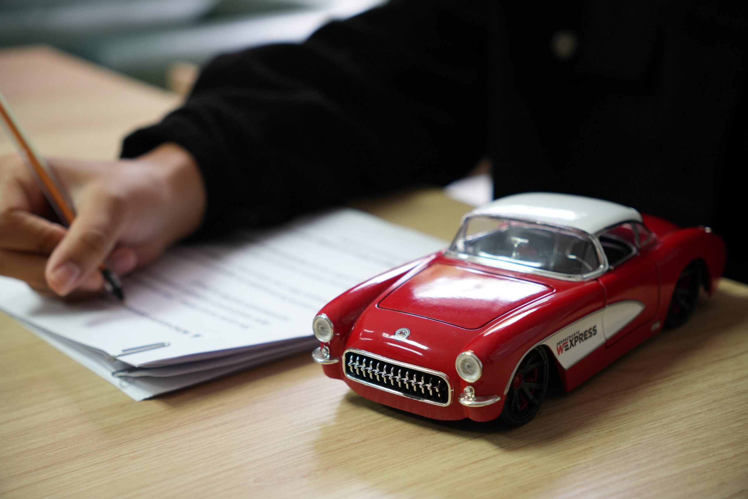 Pessoa preenchendo formulário ao lado de miniatura de carro vermelho com a logo da W Express, simbolizando o processo de liberação de veículo apreendido com documentação em mãos.
